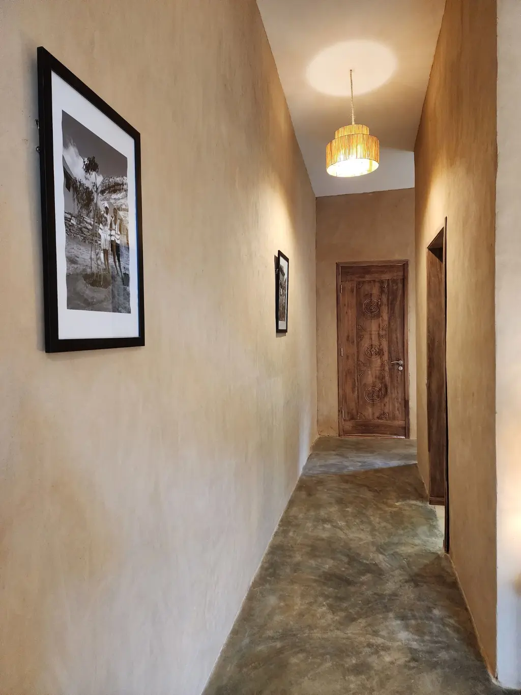Couloir et artisanat malgache dans une Suite Signature à Anja. Couloir intérieur d’une Suite Signature de The Anja Reserve Lodge à Ambalavao, avec porte sculptée en bois, suspension en raphia et photographies inspirées de Madagascar près de la Réserve d’Anja.