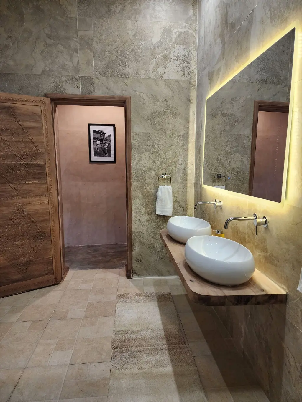 Salle de bain élégante avec double vasque à Anja. Double vasque dans la salle de bain d’une Suite Signature de The Anja Reserve Lodge à Ambalavao, avec miroir lumineux, meuble en bois et porte sculptée près de la Réserve d’Anja.